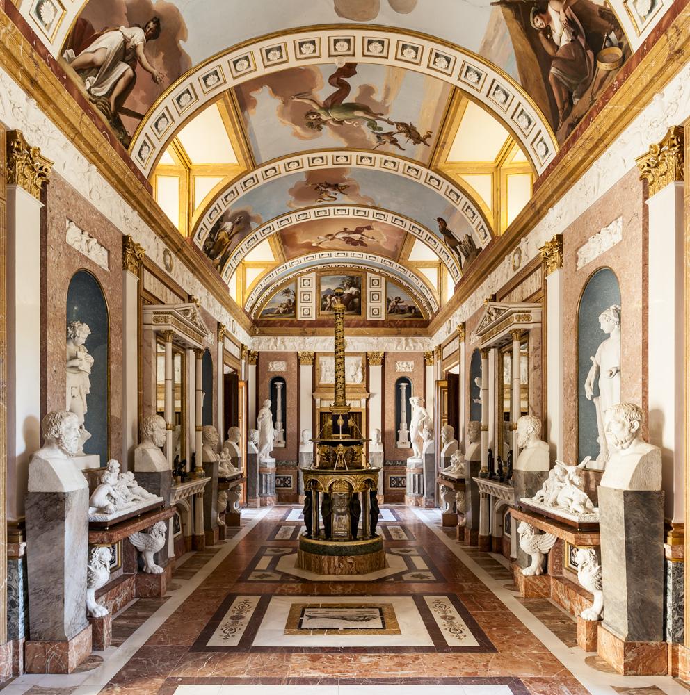 La galerie des statues présente les bustes des sages et des philosophes grecs. Sur le sol, quelques mosaïques romaines de Mérida et au centre de la pièce, une horloge unique connue sous le nom de "Colonne de Trajan" fabriquée à Paris en 1804. – © Patrimonio Nacional