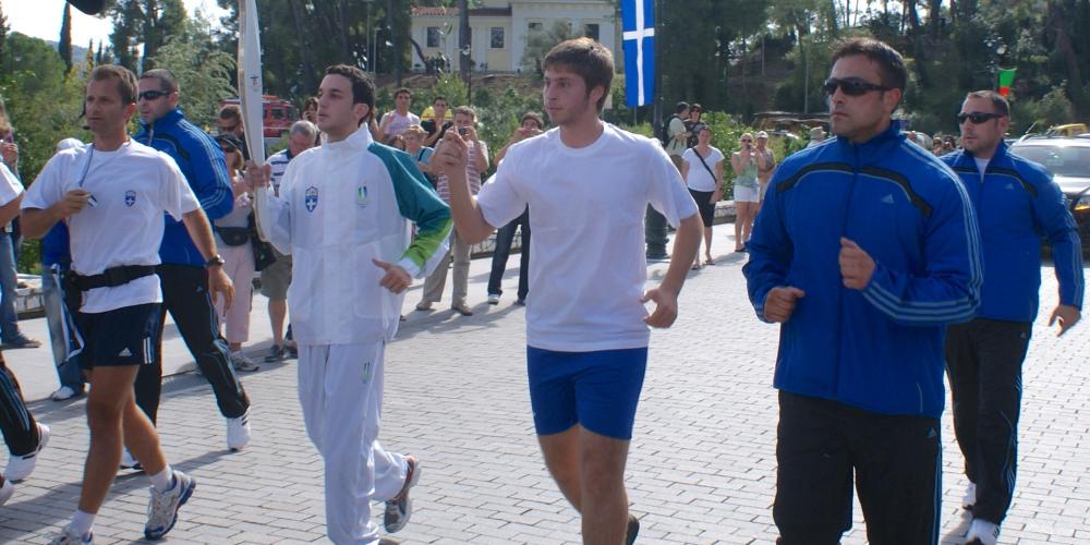 The group of runners carrying the Olympic Flame departs from the City of Olympia. The Museum of the History of the Olympic Games of Antiquity is visible in the background. – © Hellenic Ministry of Culture and Sports / Ephorate of Antiquities of Ilia