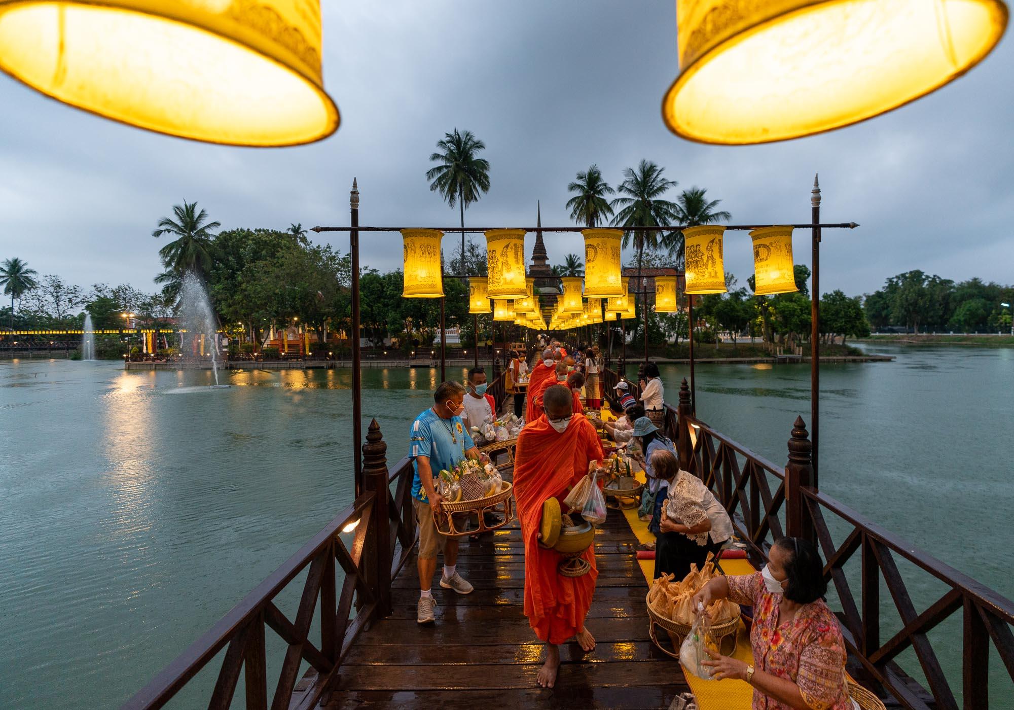 Lanterns light the bridge as the monks walk out from the temple to receive the offerings from worshippers. – © Michael Turtle