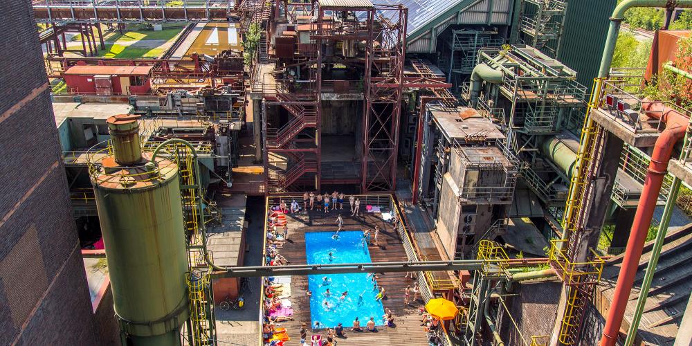 Une piscine bleu clair au milieu de la cokerie de Zollverein : Pendant les vacances d'été en Rhénanie-du-Nord-Westphalie, les visiteurs de Zollverein peuvent se rafraîchir et se détendre dans cette piscine. – © Jochen Tack / Zollverein Foundation