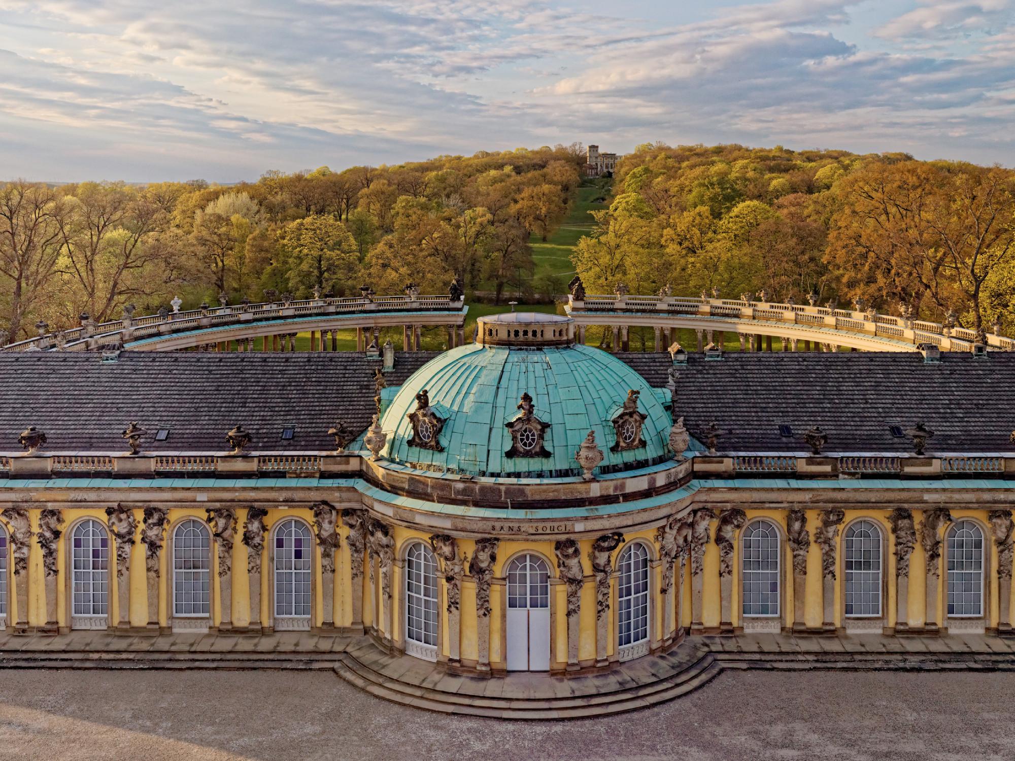 In contrast with the playful design of the garden façade, the north façade is majestic and commanding. A double row of eighty-eight Corinthian columns forms two colonnades in quarter-circle segments enclosing a semicircular cour d'honneur. The Ruins Hill is aligned with this axis to the north.  - © A. Stiebitz / SPSG