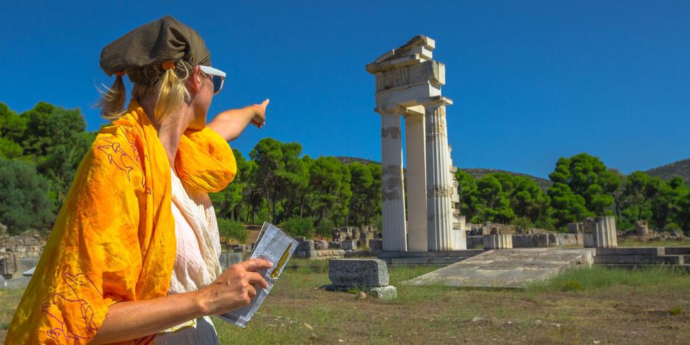 Tourist woman in Greek dress indicates the ruins of Temple of Asklepieion, Epidaurus, Peloponnese, Greece. The Sanctuary of Asclepius is a famous heritage site. – © Benny Marty / Shutterstock Tourist woman in Greek dress indicates the ruins of Temple of Asklepieion, Epidaurus, Peloponnese, Greece. The Sanctuary of Asclepius is a famous heritage site. – © Benny Marty / Shutterstock