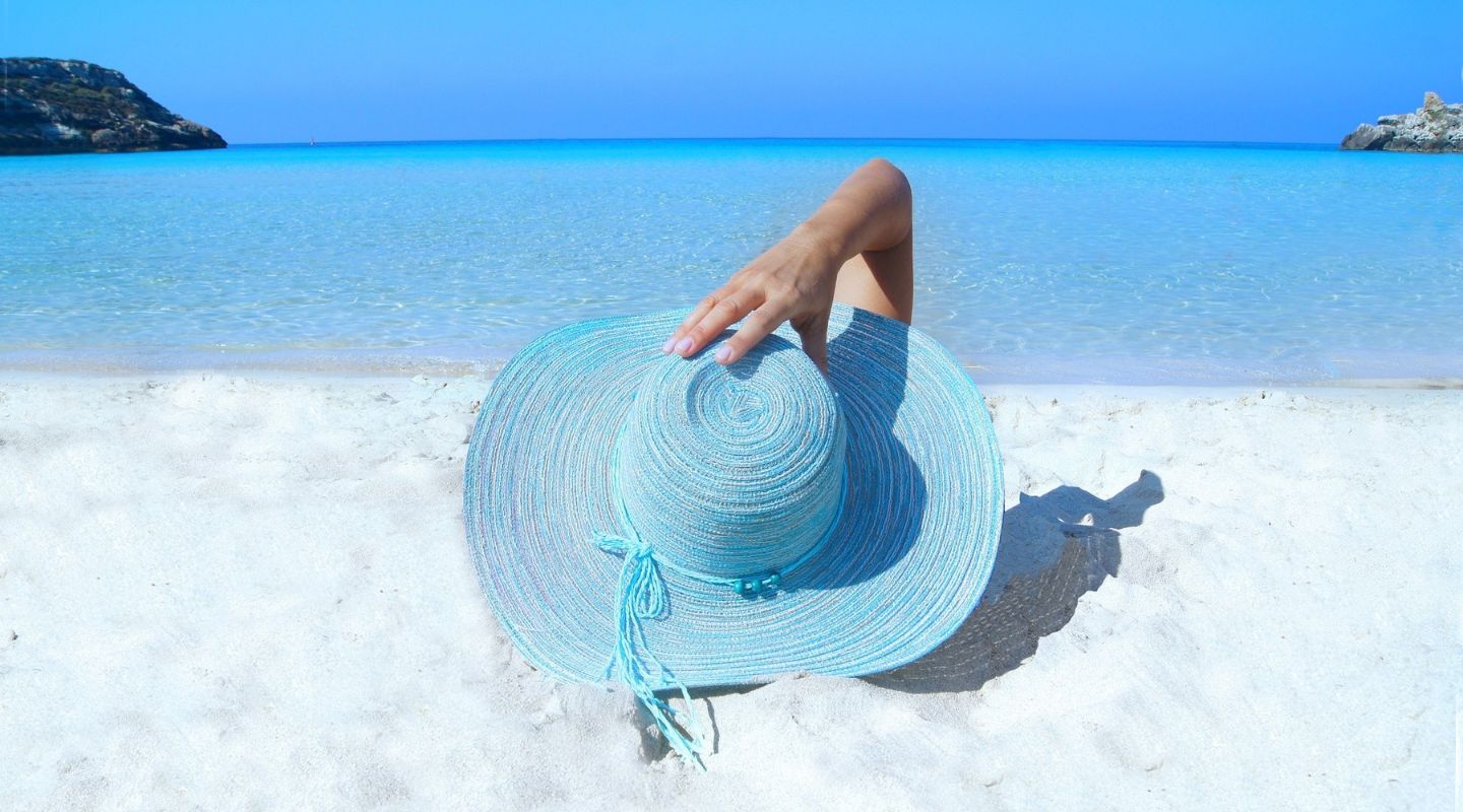 A woman lays on white sands, holding a large blue hat over her head