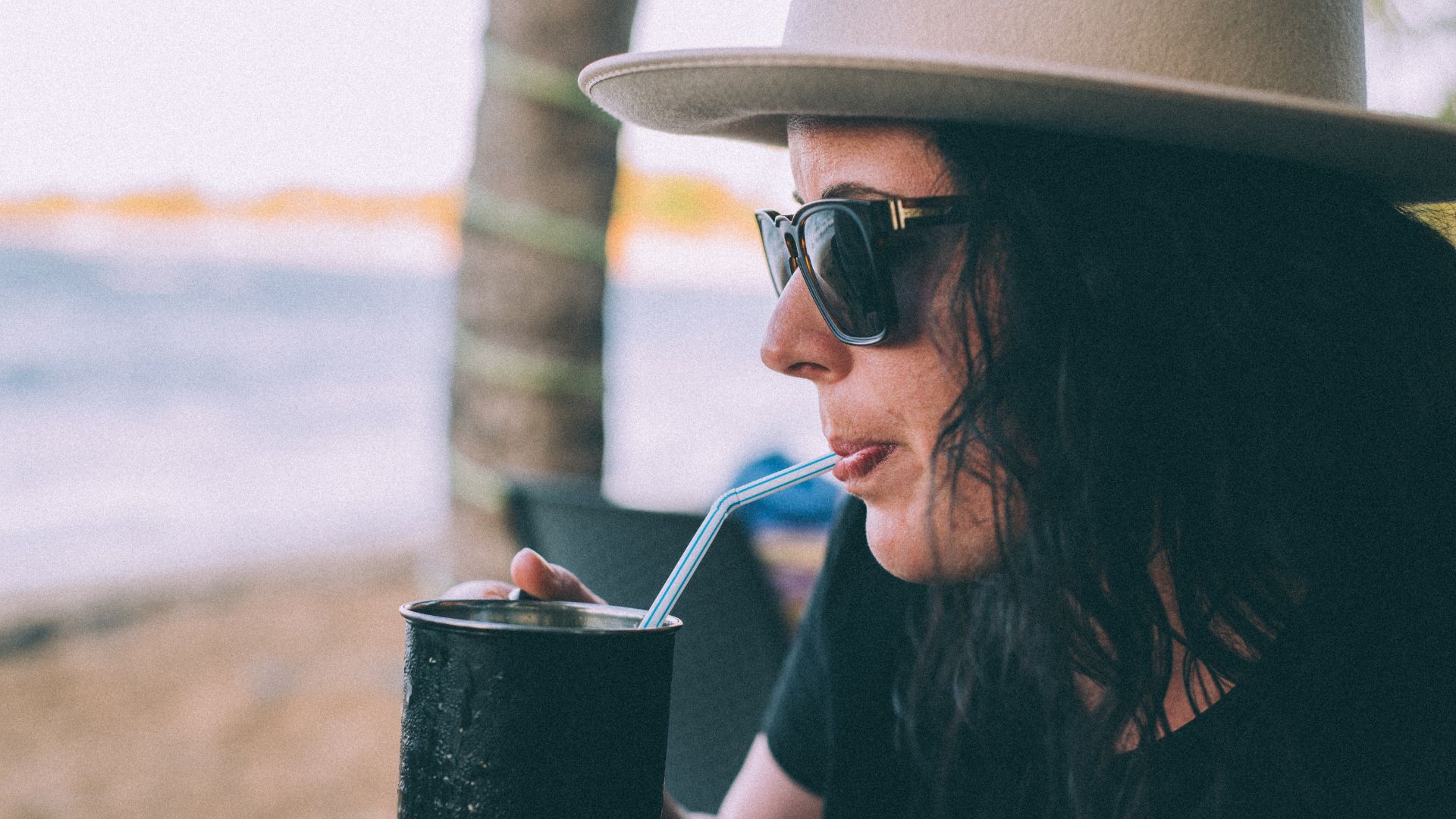 Close-up shot of a woman dressed in beach-attire, drinking from a straw, looking at the horizon. 