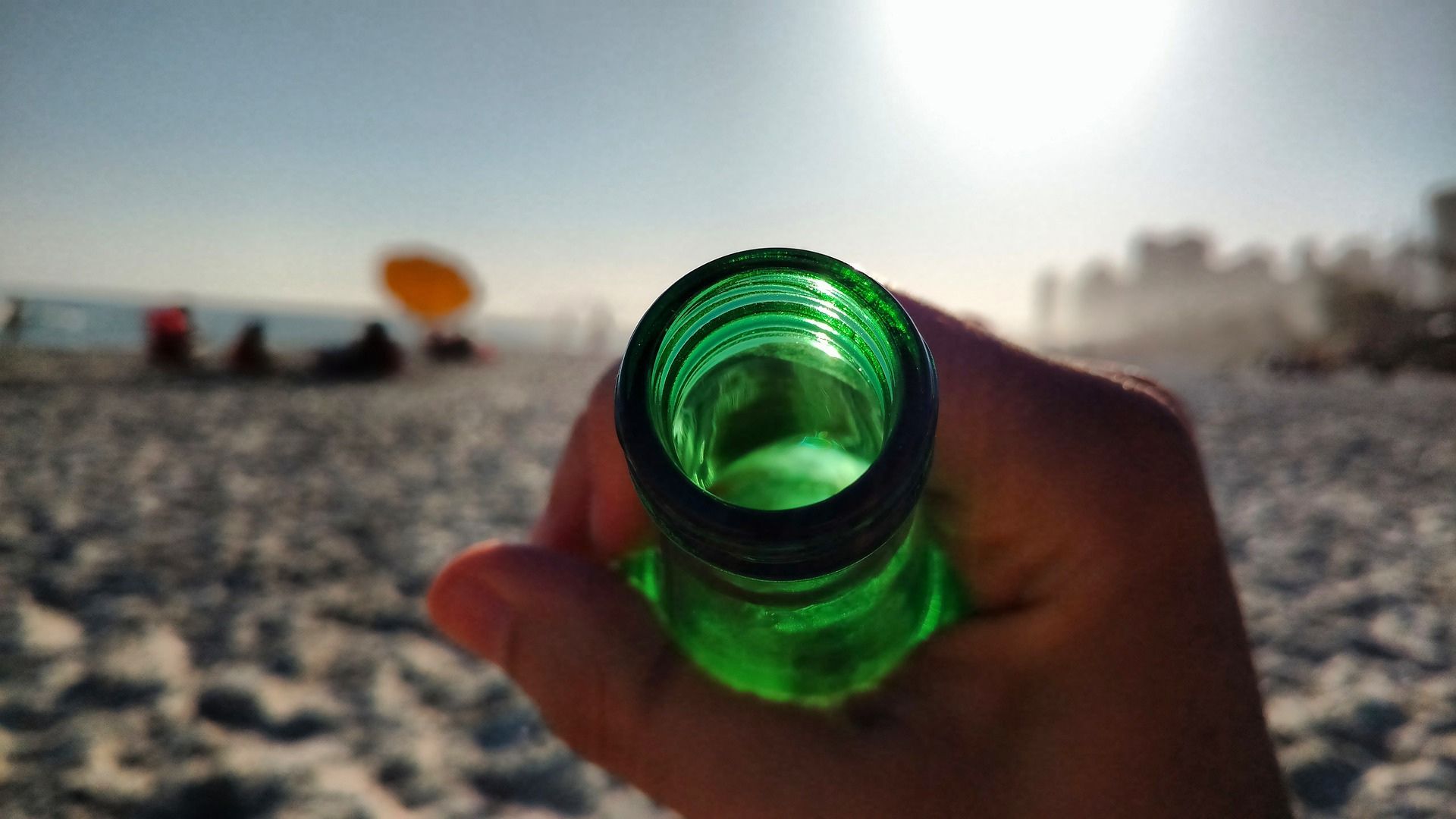 Photo of a green bottle being held by someone's hand. That someone is laying on the beach.