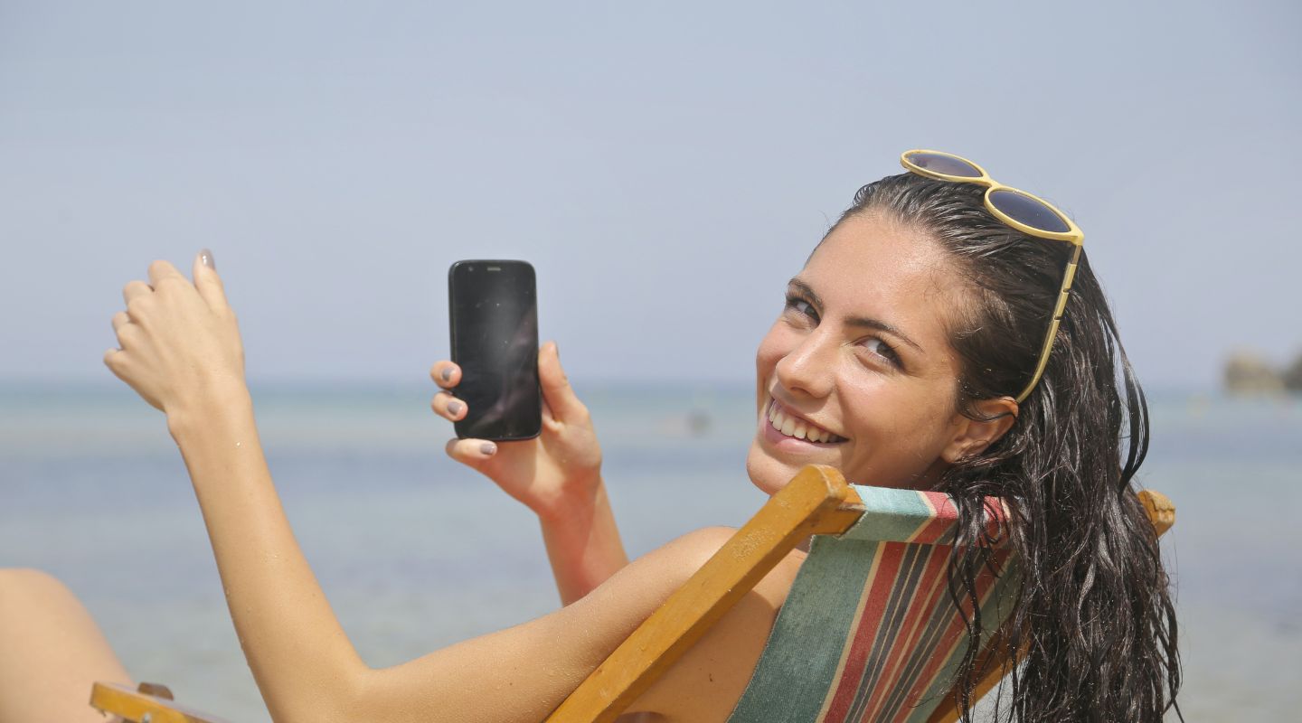 A woman on the beach holds her phone and looks at the camera