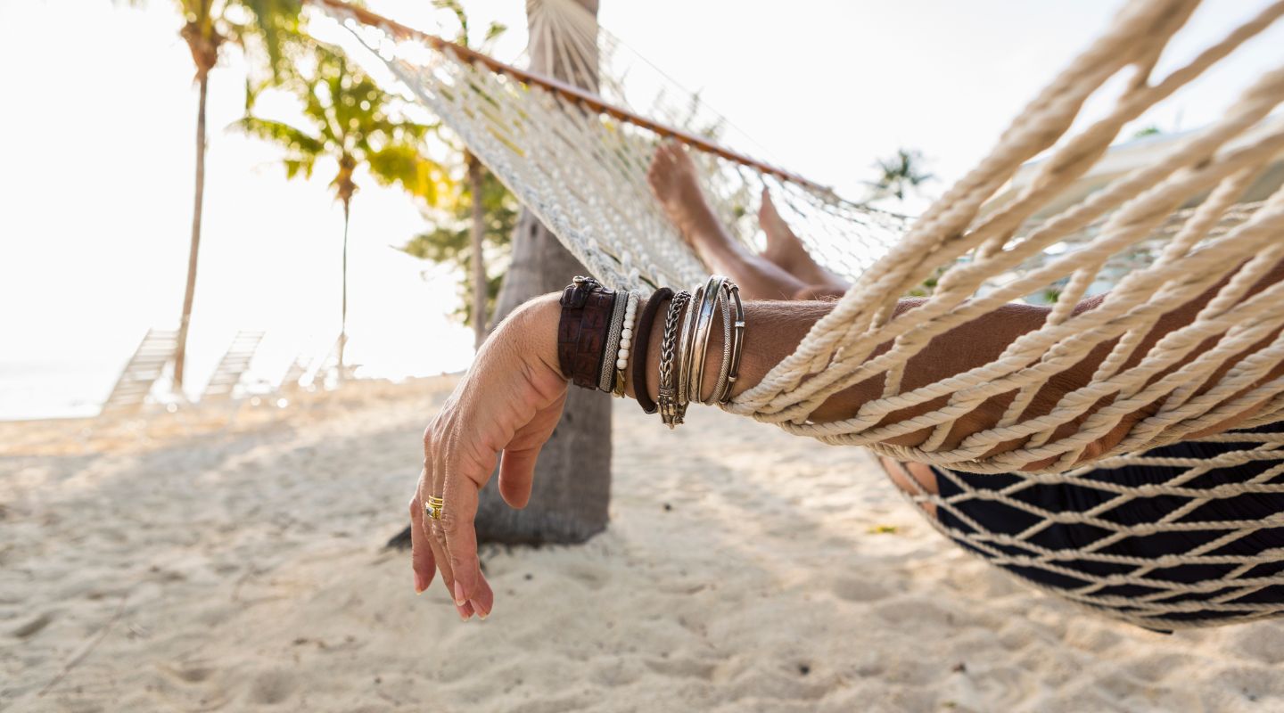A bejeweled arm hangs from the side of a hammock hanging in front of the ocean.