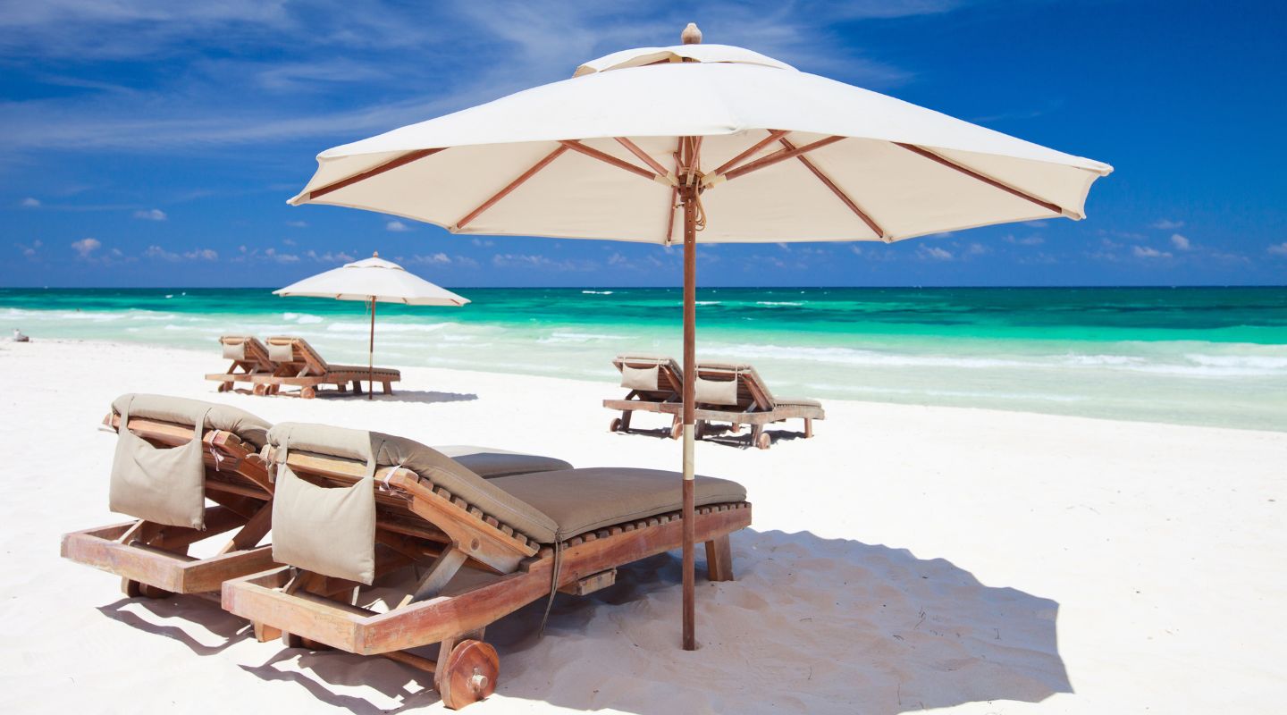 Comfortable-looking beach chairs under parasols, placed on white sand, with the ocean at the background. 