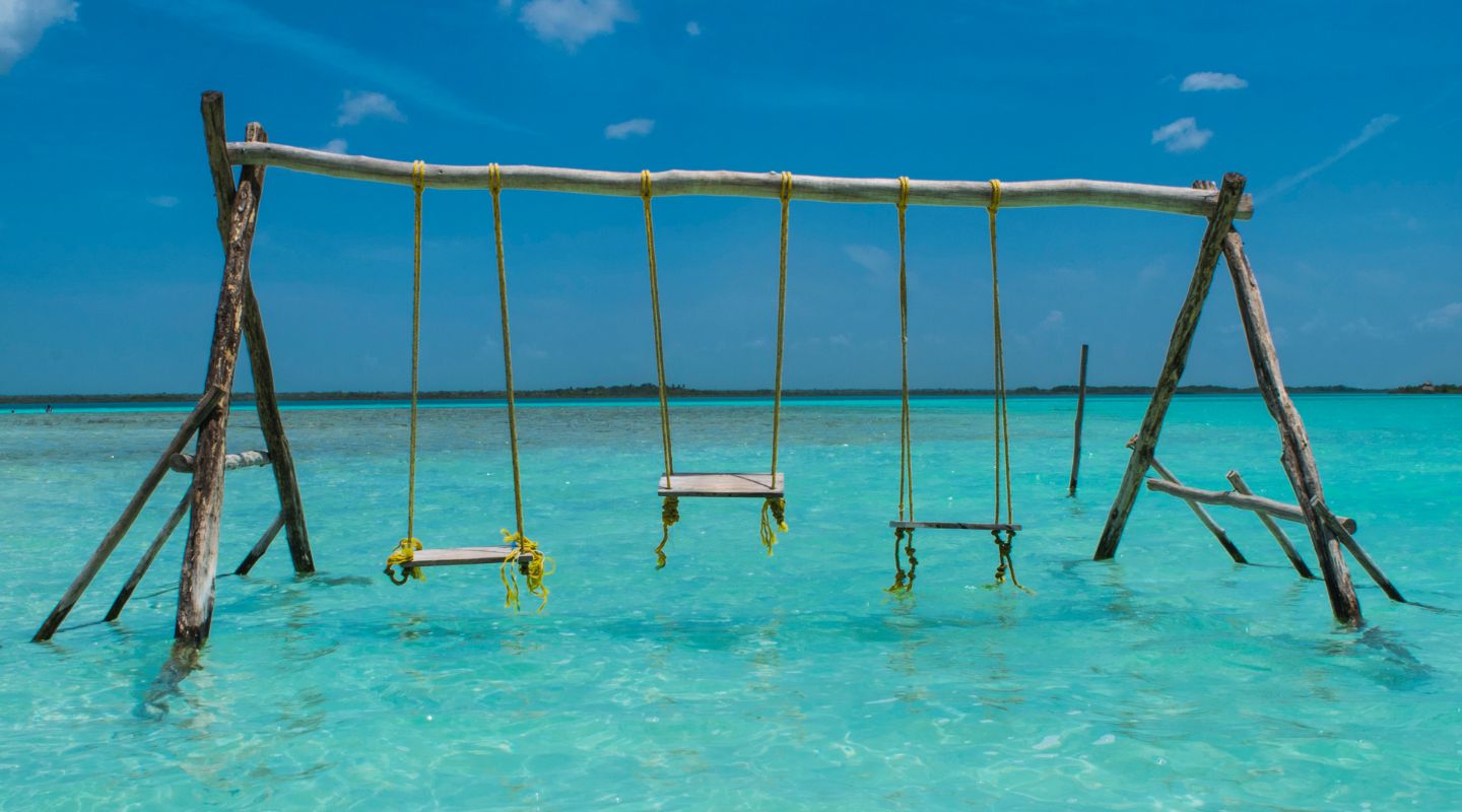 A trio of rustic-looking swings hang above the blue waters of Bacalar
