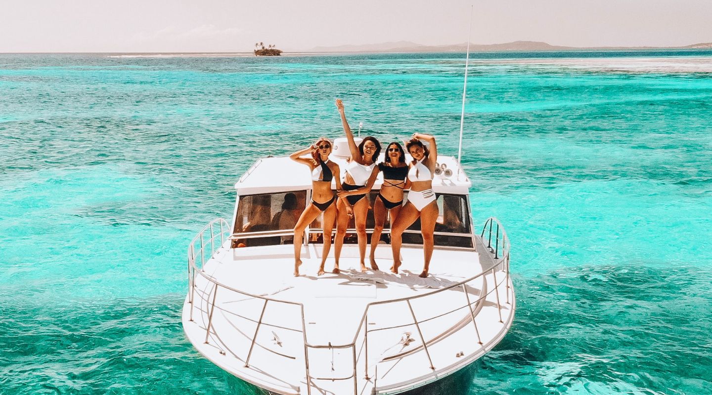 A group of women stand atop a boat as they smile and wave at the camera