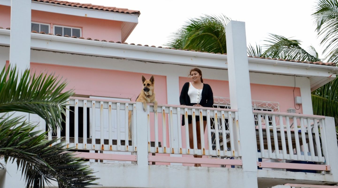 A woman and a dog stand on a house's balcony. The house is painted a light pink.