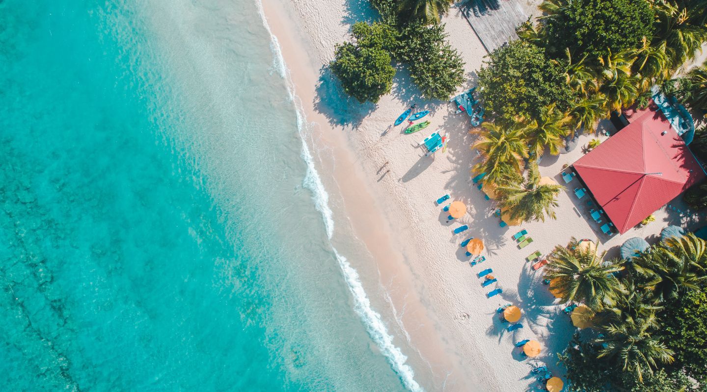 Overhead view of the beach, with a small red-roofed shack built on the sand and plenty of beach chairs and umbrellas facing the ocean.