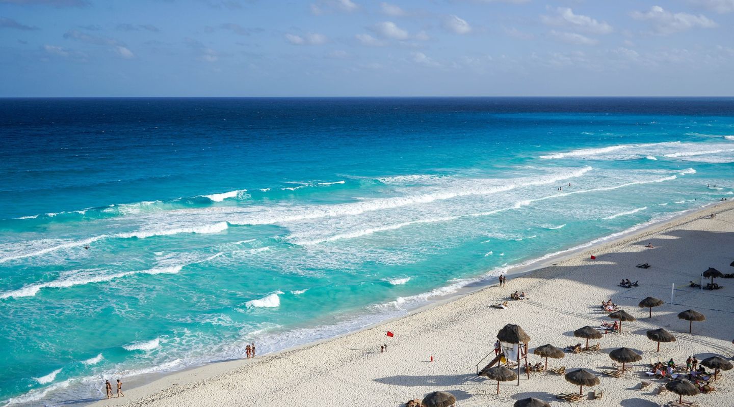 Photo of the beach, with the ocean on the left and the beach on the right. There are sun loungers and straw umbrellas  on the sand.