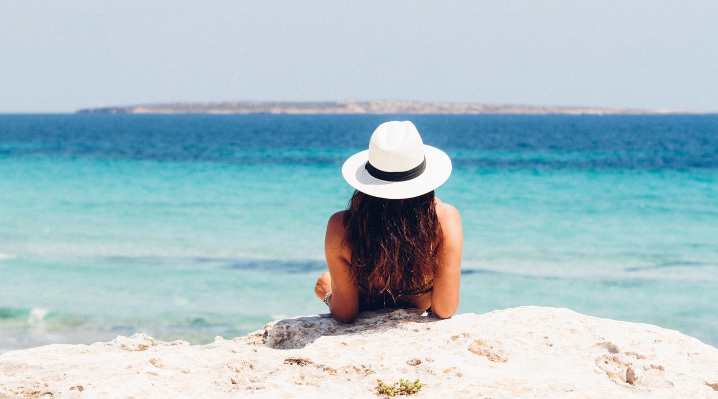 Woman's upper body seen from behind. She's wearing a large hat and is looking at the ocean.