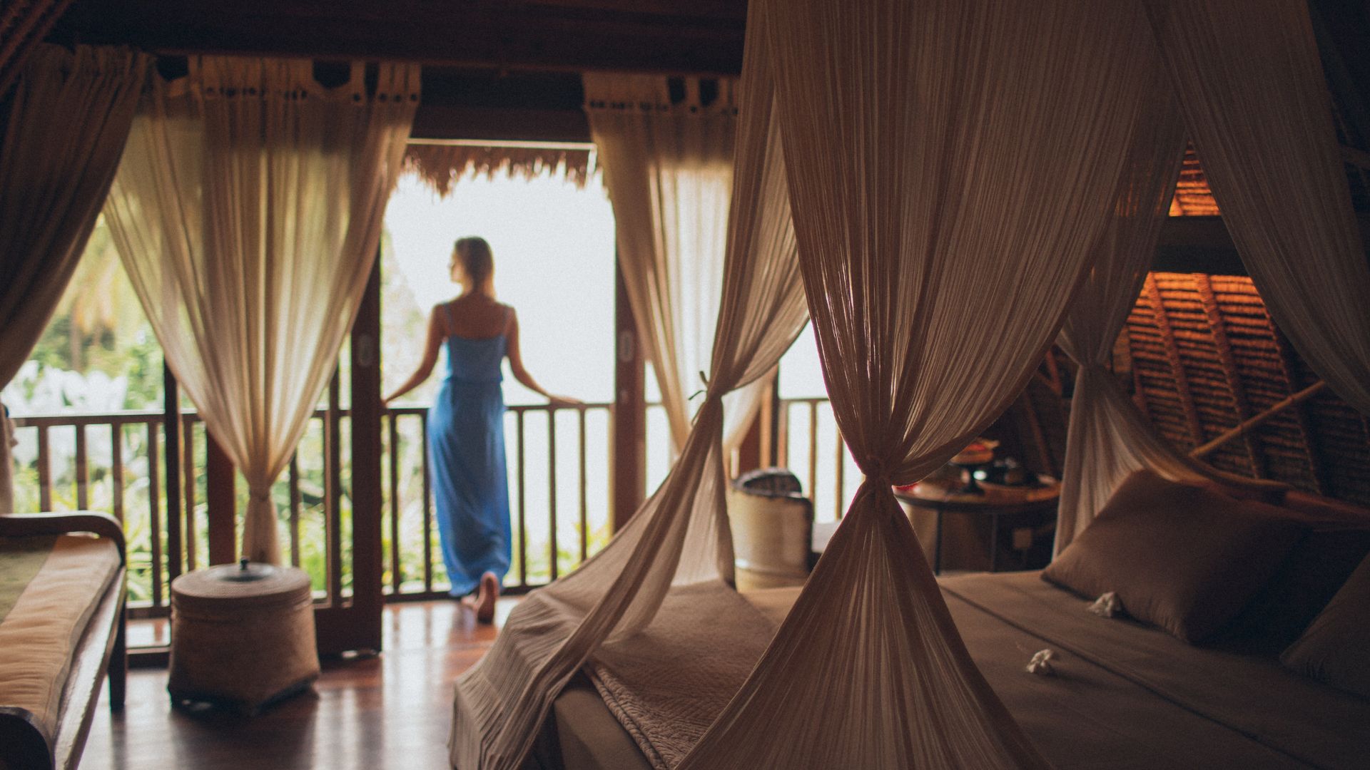 A woman wearing a blue dress has her back to the camera as she stands on the balcony of what appears to be a beach hotel room